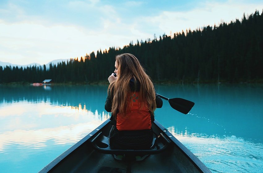 Person in a red life jacket paddling a canoe on a calm turquoise lake with forested hills in the background at sunset/dusk