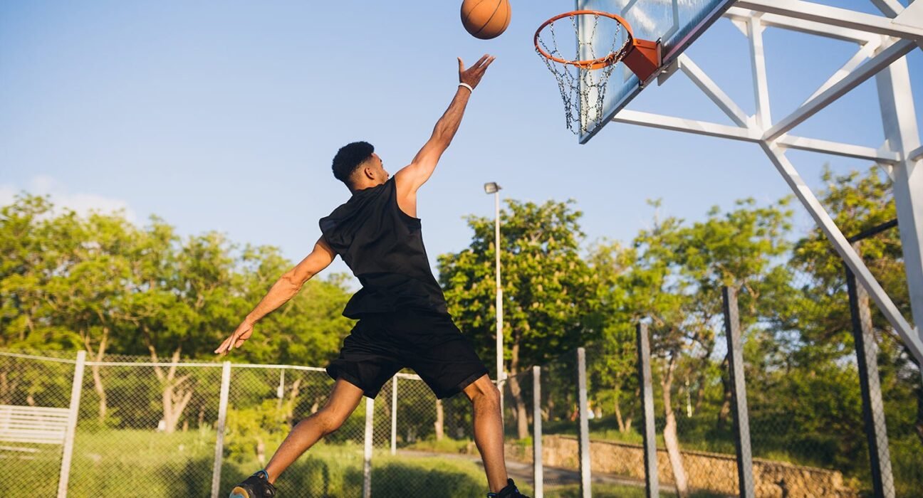 find more africa #findmoreafrica › Player in black jumps to dunk a basketball on an outdoor court under a clear blue sky.