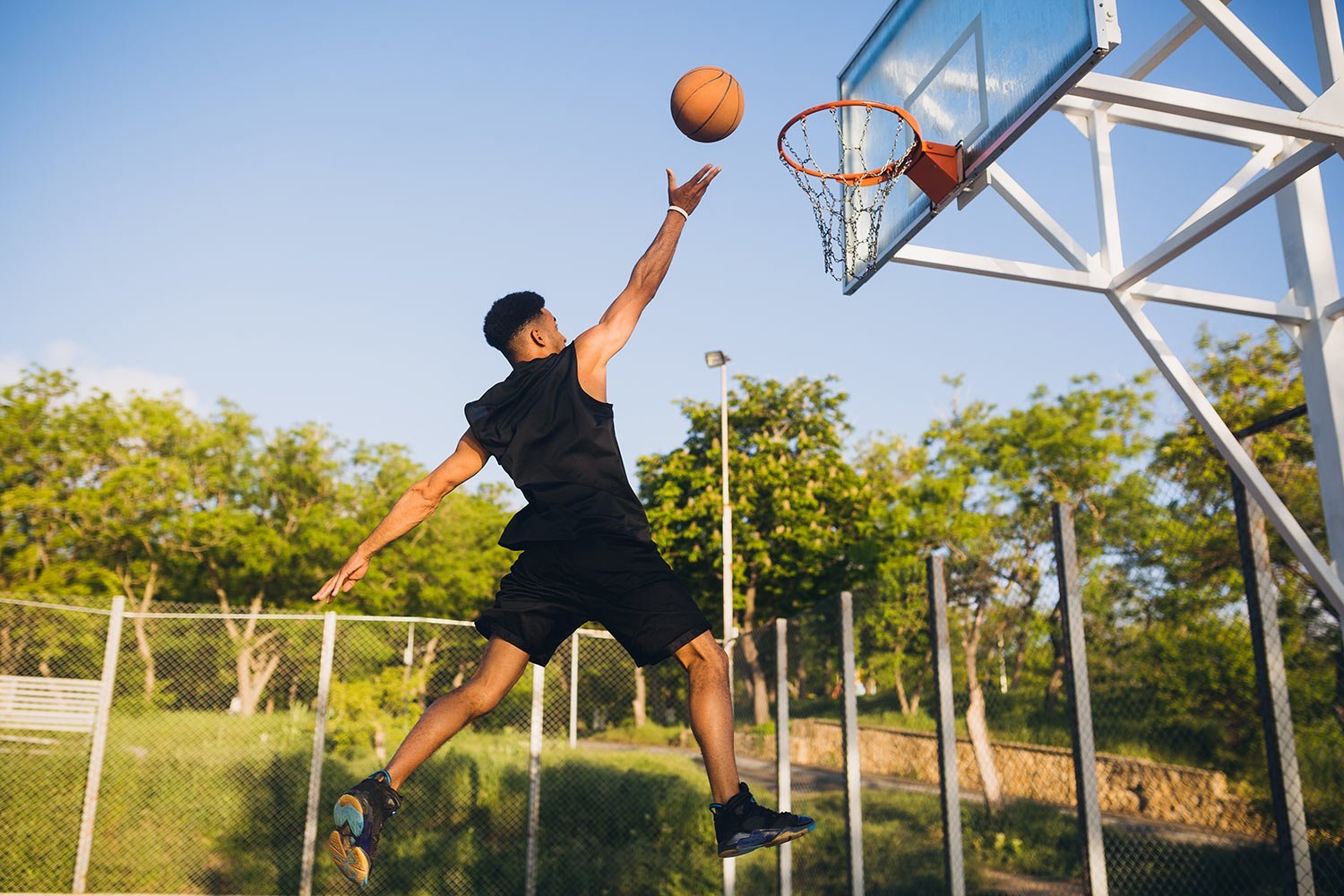 Player in black jumps to dunk a basketball on an outdoor court under a clear blue sky.