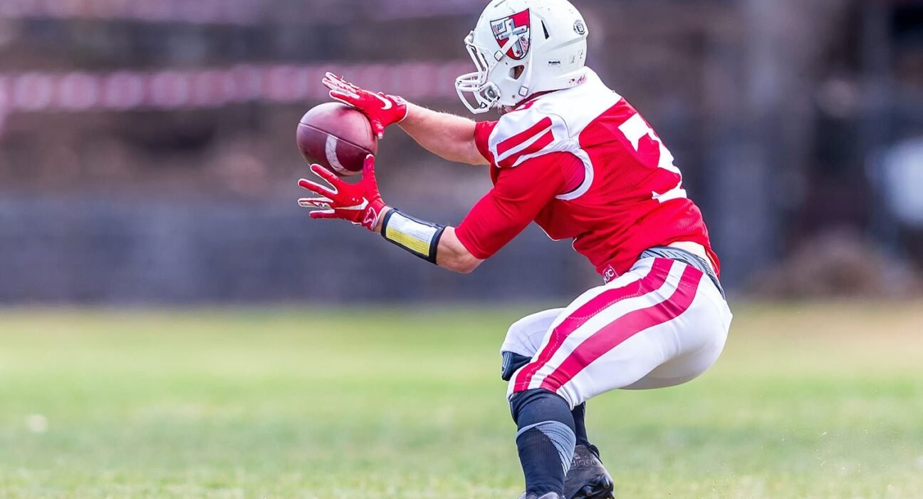 American football player in red and white uniform dives to catch a football on a grassy field, wearing a white helmet and red gloves.