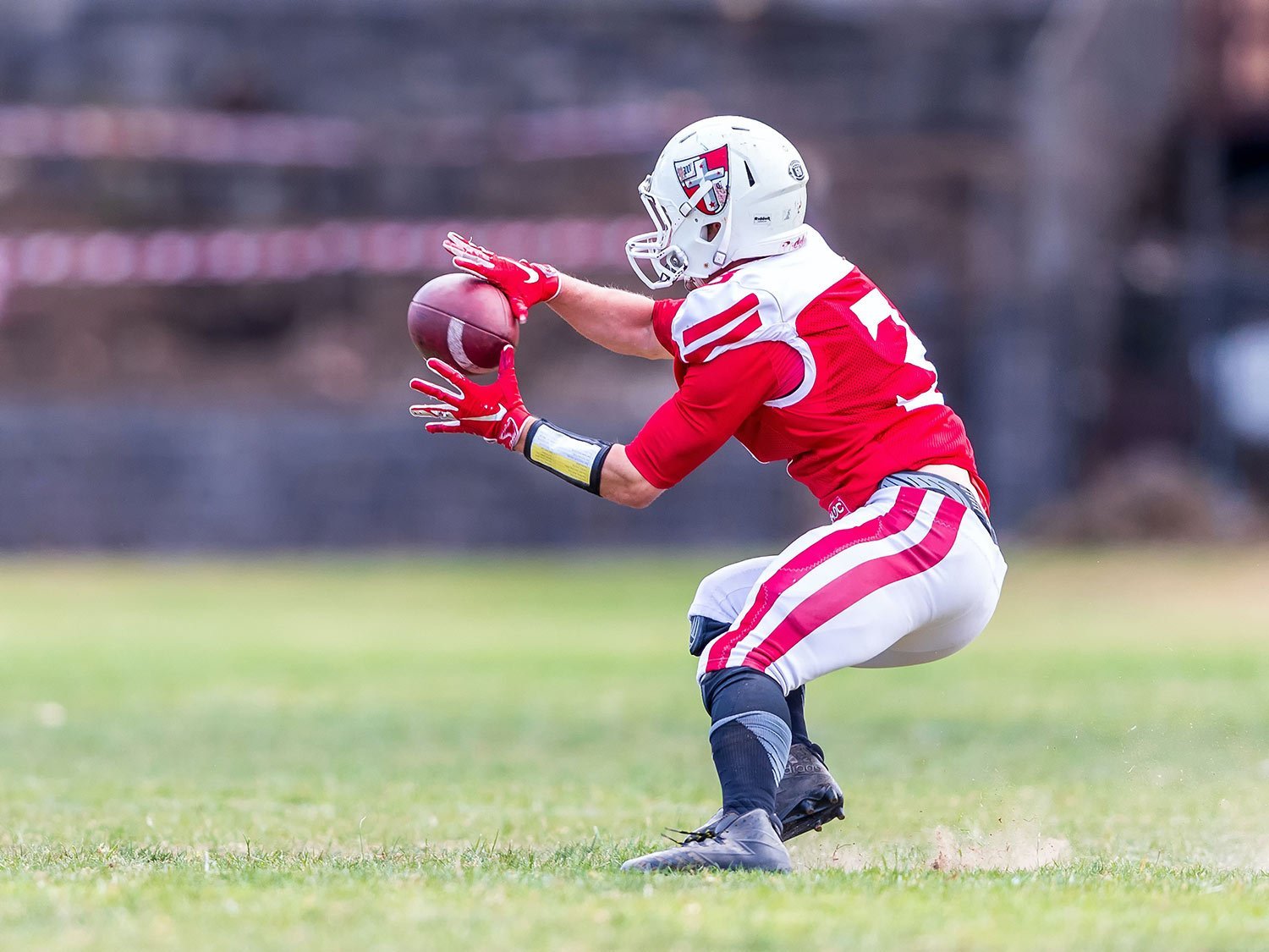 American football player in red and white uniform dives to catch a football on a grassy field, wearing a white helmet and red gloves.