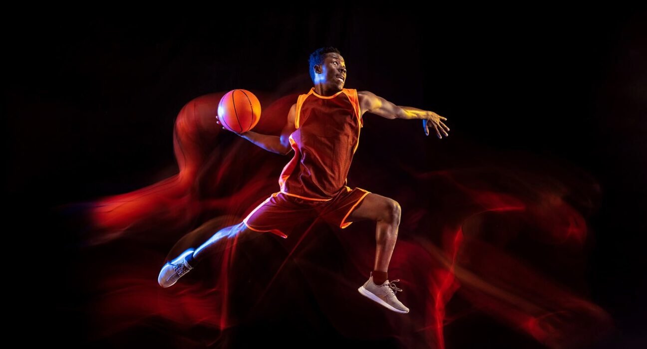 Basketball player in an orange uniform jumping mid-air with the ball in hand against a dark background with red light trails.