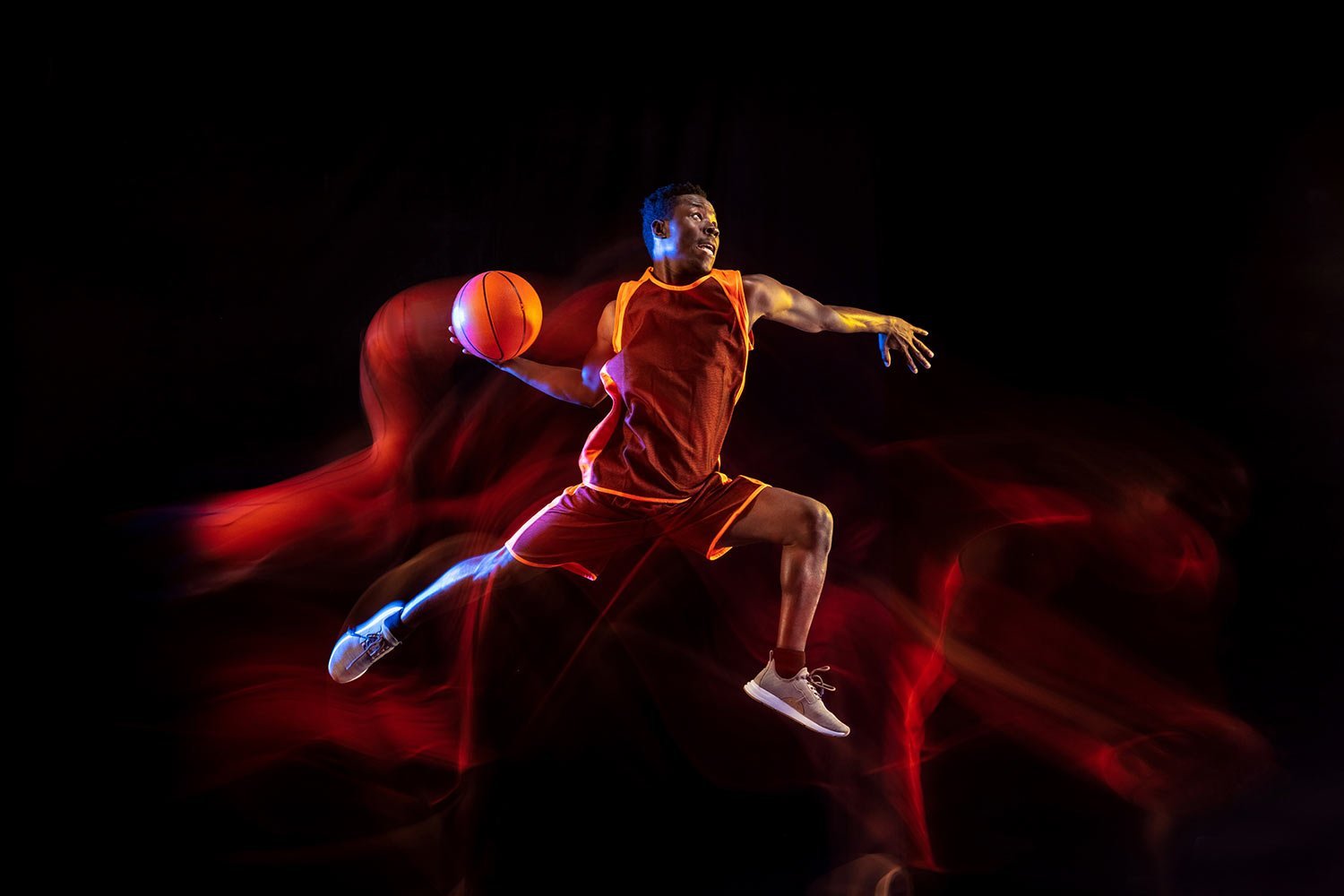 Basketball player in an orange uniform jumping mid-air with the ball in hand against a dark background with red light trails.