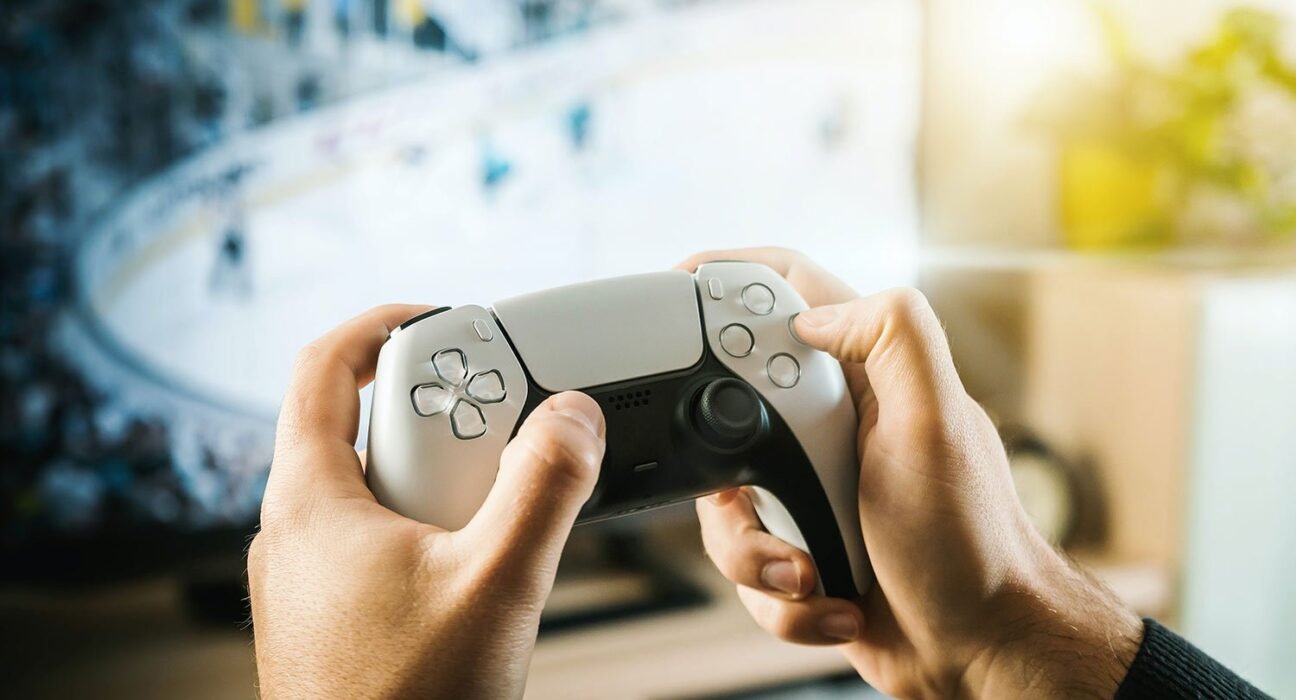 Person holds a white PlayStation 5 DualSense controller, gaming on a blurred screen in the background.