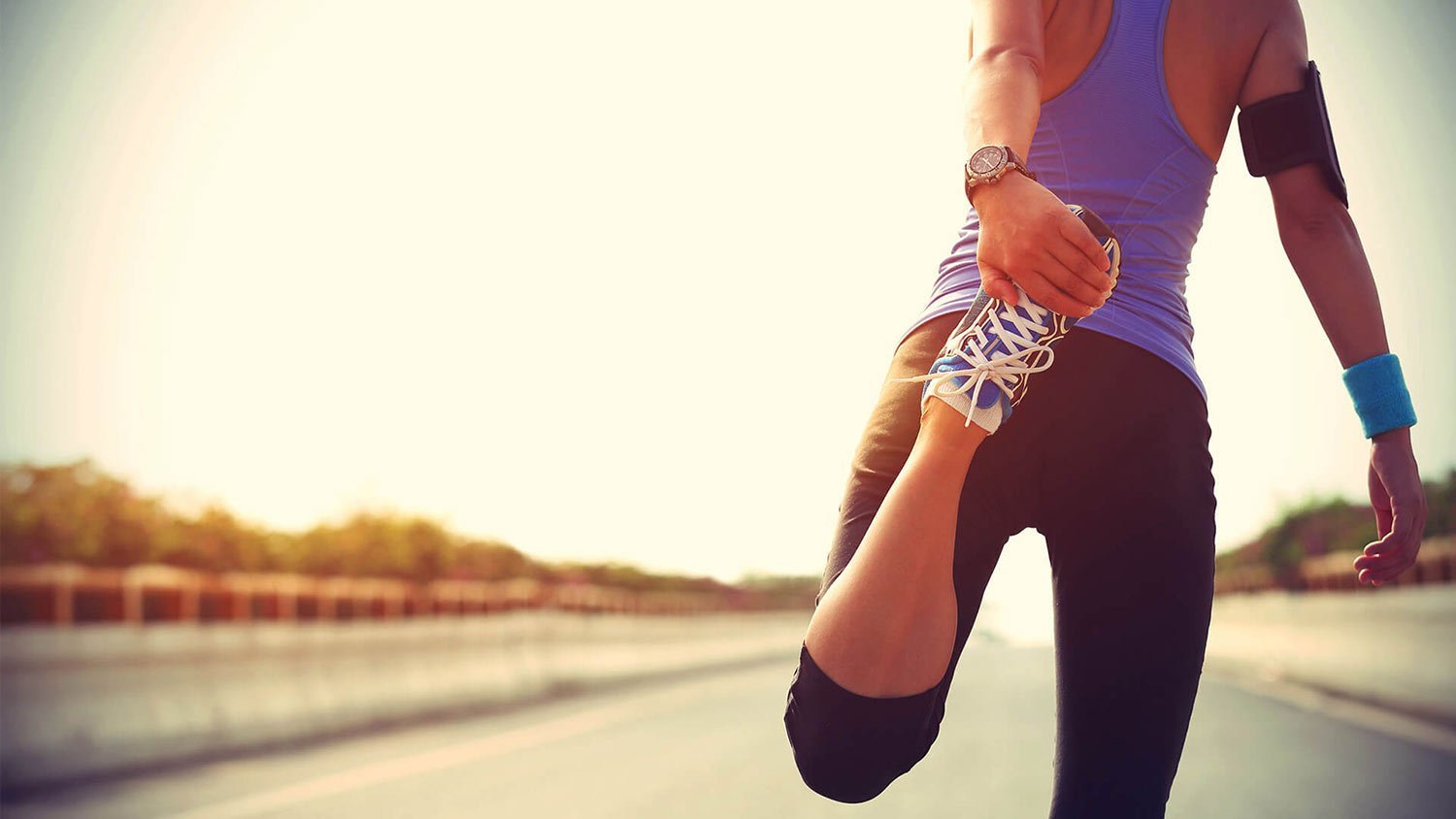 Runner on an open road ties a blue-and-white sneaker, preparing for a run at sunset.