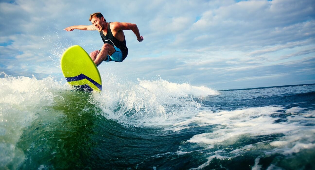 Male surfer riding a neon yellow-green board on a breaking blue-green wave under a cloudy sky.