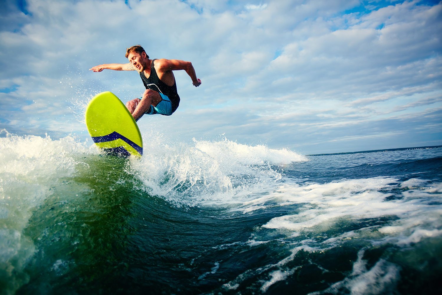 Male surfer riding a neon yellow-green board on a breaking blue-green wave under a cloudy sky.