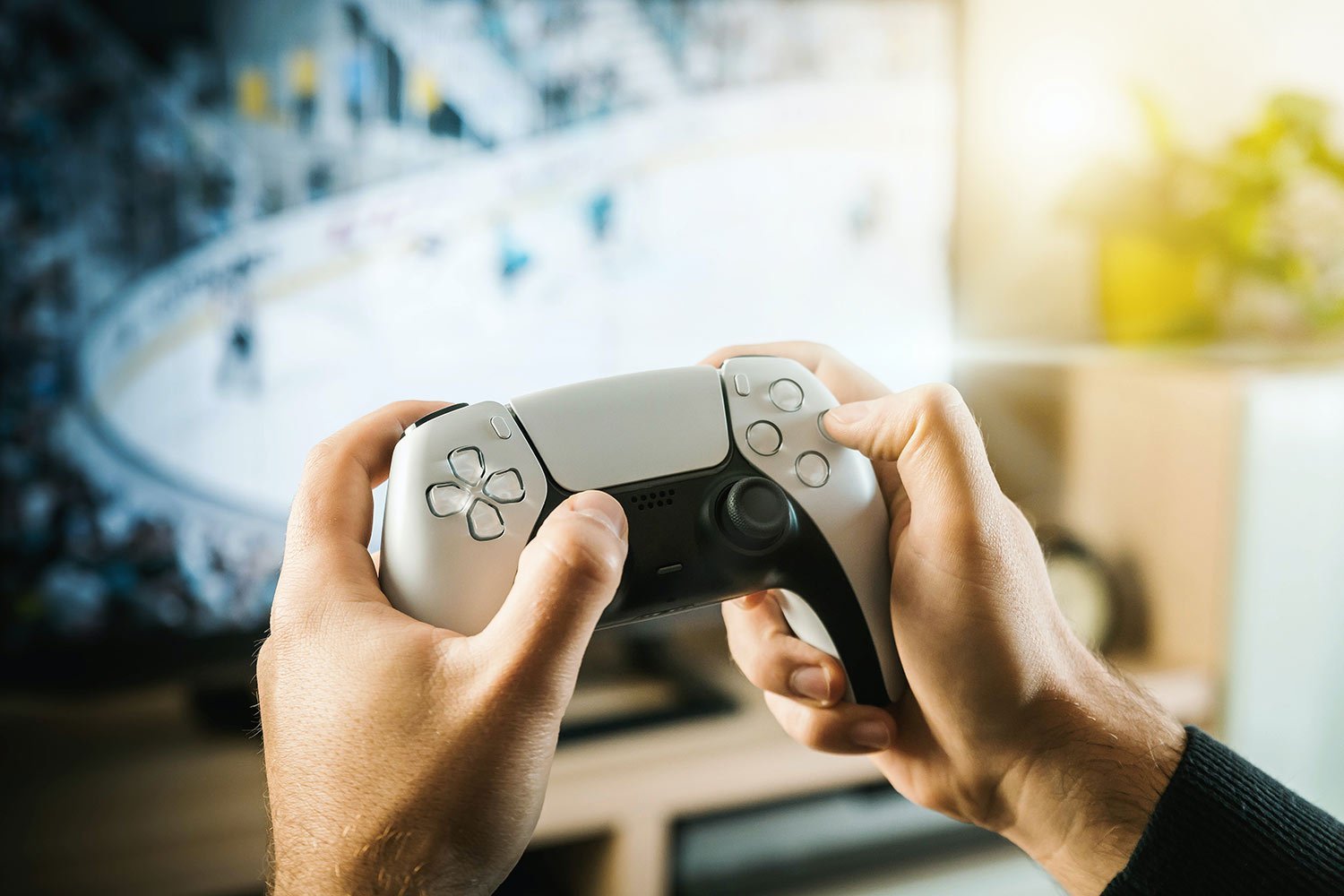 Person holds a white PlayStation 5 DualSense controller, gaming on a blurred screen in the background.