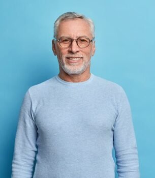 Smiling older man with gray hair and glasses wearing a light blue knit sweater against a blue backdrop.