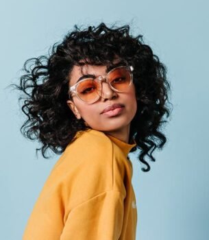Portrait of a person with curly hair wearing orange-tinted sunglasses and a yellow sweatshirt against a light blue background.