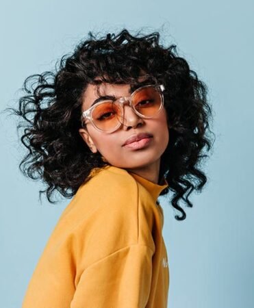 Portrait of a person with curly hair wearing orange-tinted sunglasses and a yellow sweatshirt against a light blue background.