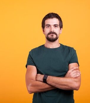 Bearded man in a teal t-shirt with arms crossed, standing against a bright orange background looking at the camera.
