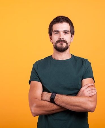 find more africa #findmoreafrica › Bearded man in a teal t-shirt with arms crossed, standing against a bright orange background looking at the camera.