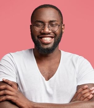 Smiling man with round glasses and a beard, arms crossed, wearing a white t-shirt against a pink backdrop.