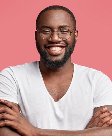 find more africa #findmoreafrica › Smiling man with round glasses and a beard, arms crossed, wearing a white t-shirt against a pink backdrop.