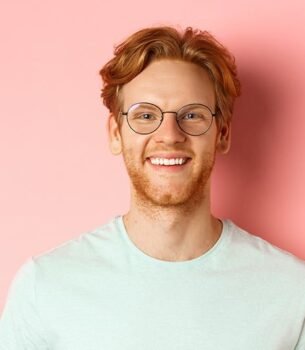 Smiling man with red hair and round glasses in a light blue T-shirt against a pink background, facing the camera.