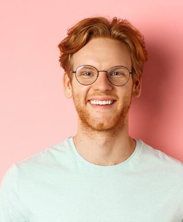 find more africa #findmoreafrica › Smiling man with red hair and round glasses in a light blue T-shirt against a pink background, facing the camera.
