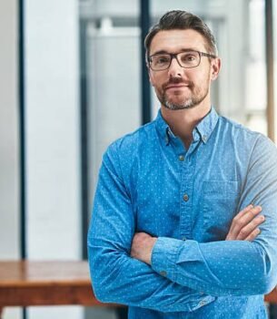 Man with glasses in a blue button-down shirt stands with arms crossed in a bright, modern office.