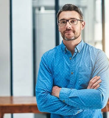 Man with glasses in a blue button-down shirt stands with arms crossed in a bright, modern office.