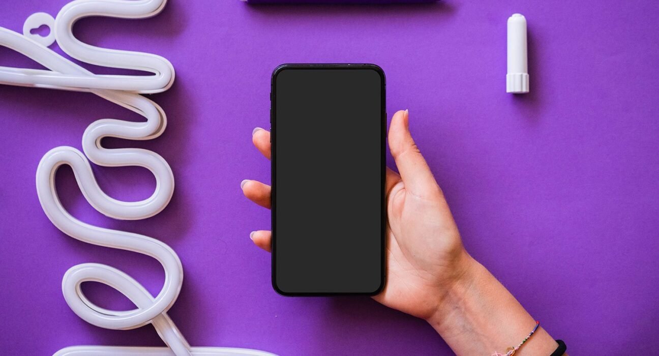 find more africa #findmoreafrica › Person holding a black smartphone with blank screen against a purple desk, surrounded by white neon tubing and a purple marker cap and pen cap on the surface