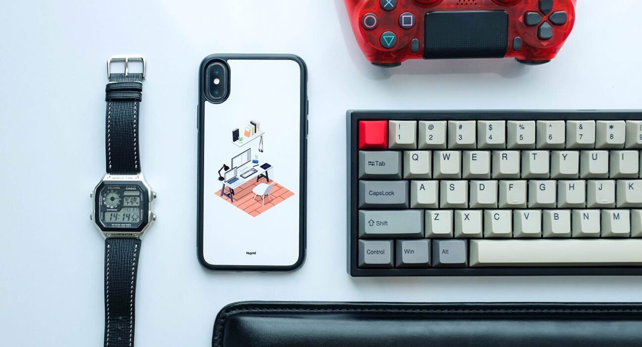 Flat lay of a tech desk: smartwatch, iPhone with illustration case, wired keyboard, red game controller, and a black wallet on a light surface