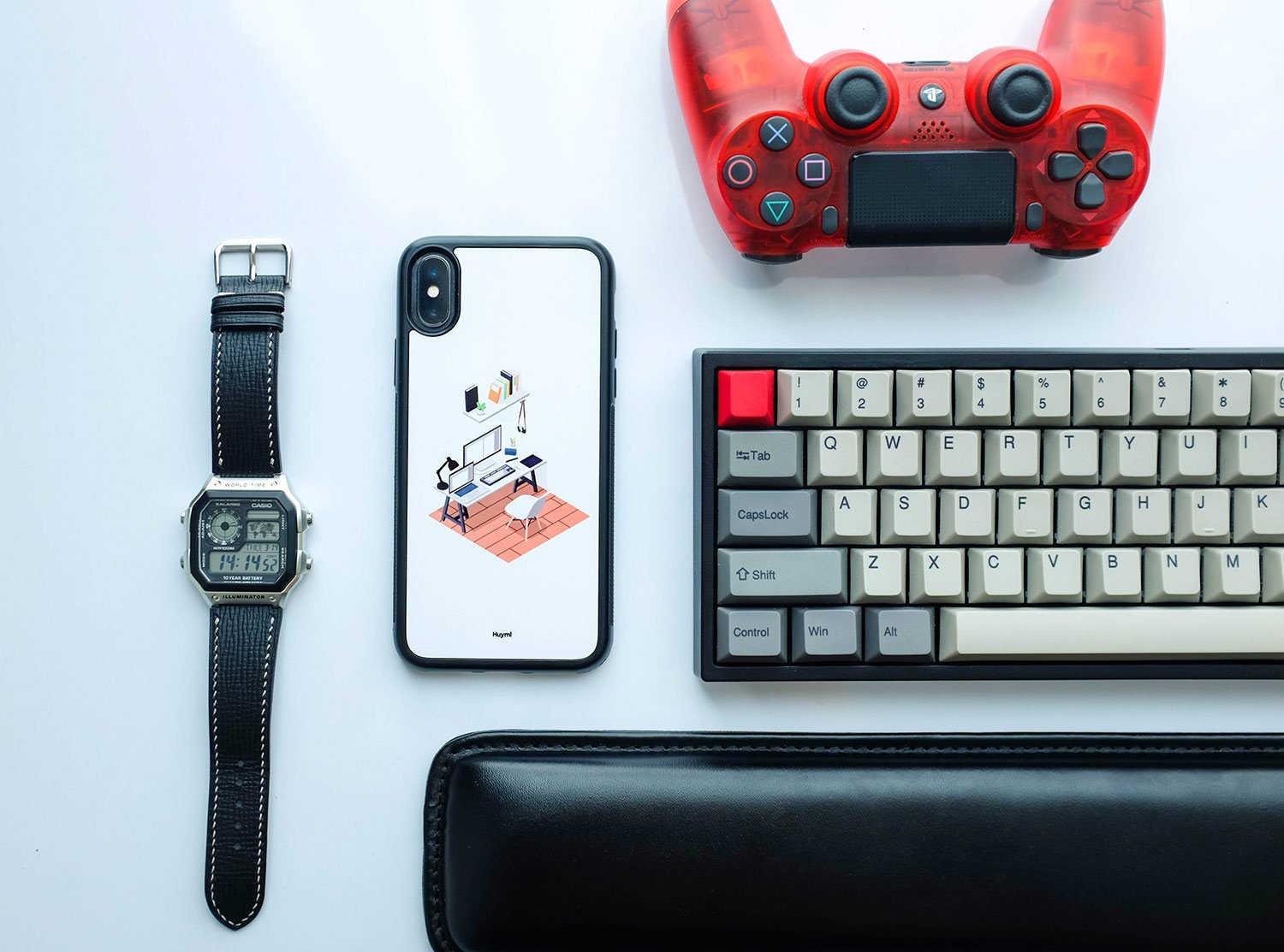 Flat lay of a tech desk: smartwatch, iPhone with illustration case, wired keyboard, red game controller, and a black wallet on a light surface