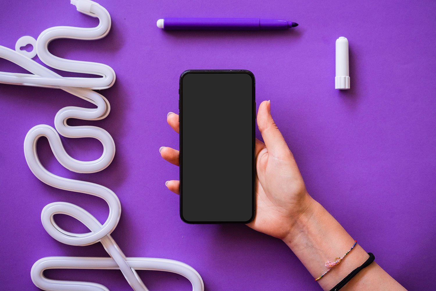Person holding a black smartphone with blank screen against a purple desk, surrounded by white neon tubing and a purple marker cap and pen cap on the surface