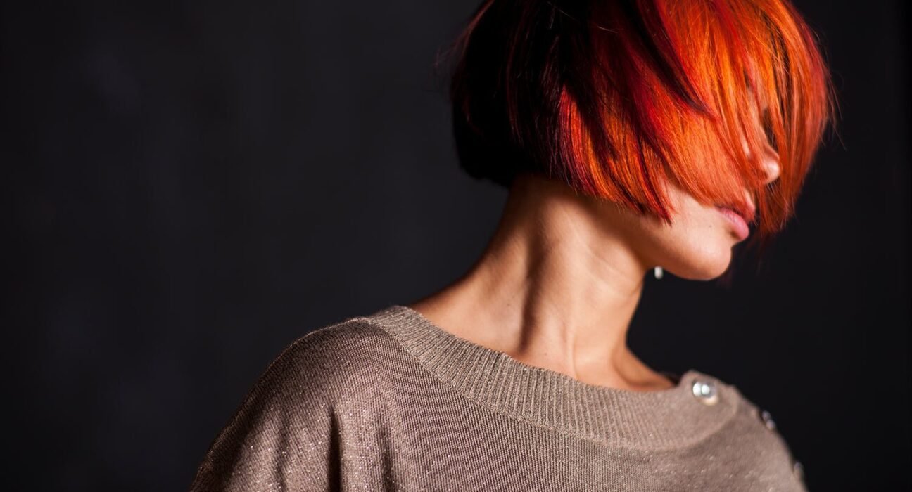 find more africa #findmoreafrica › Person with bright orange-red hair covering their face, wearing a light sparkling beige sweater, against a dark background.