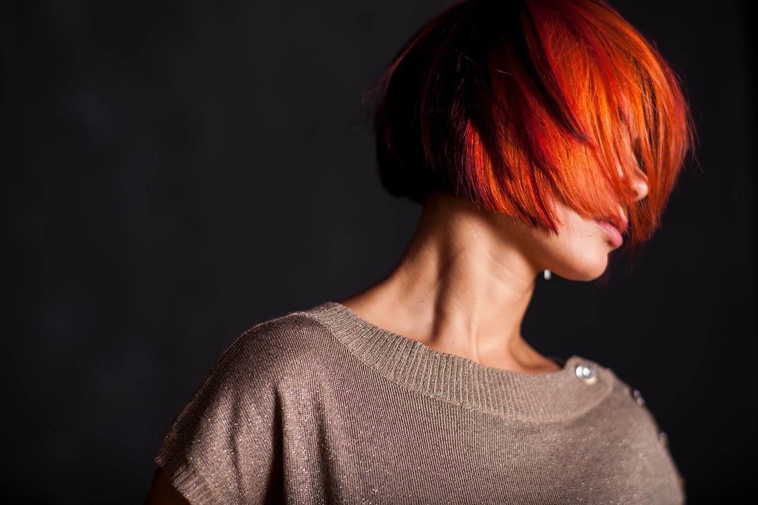 Person with bright orange-red hair covering their face, wearing a light sparkling beige sweater, against a dark background.