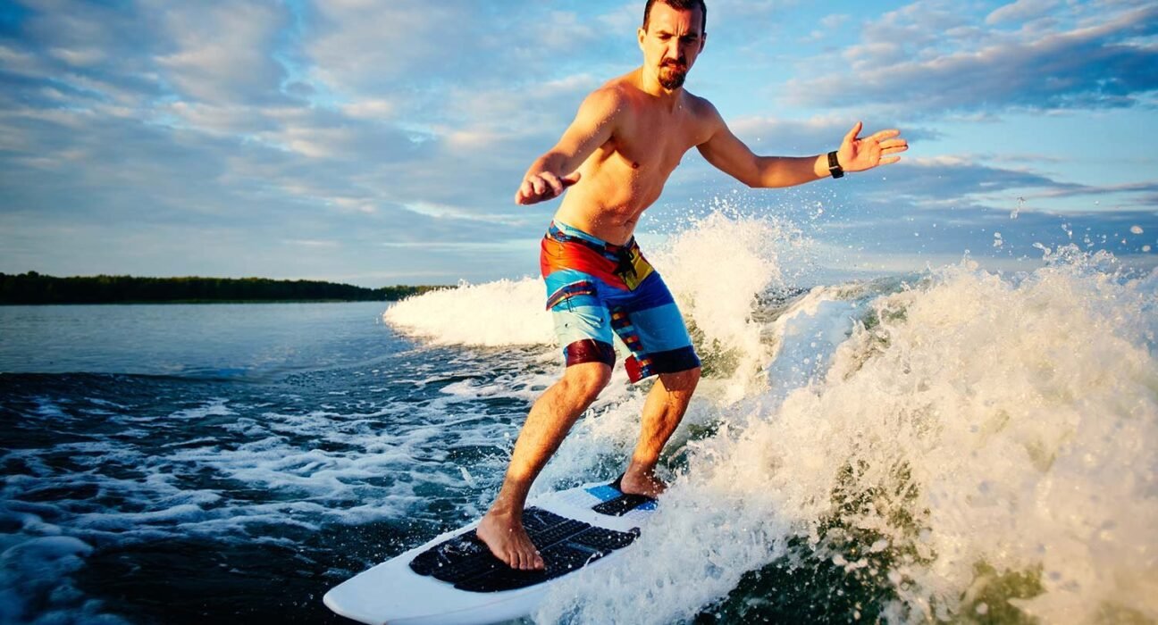 Man shirtless, wearing colorful board shorts, stands on a surfboard riding a wave with spray around him on a blue sky day