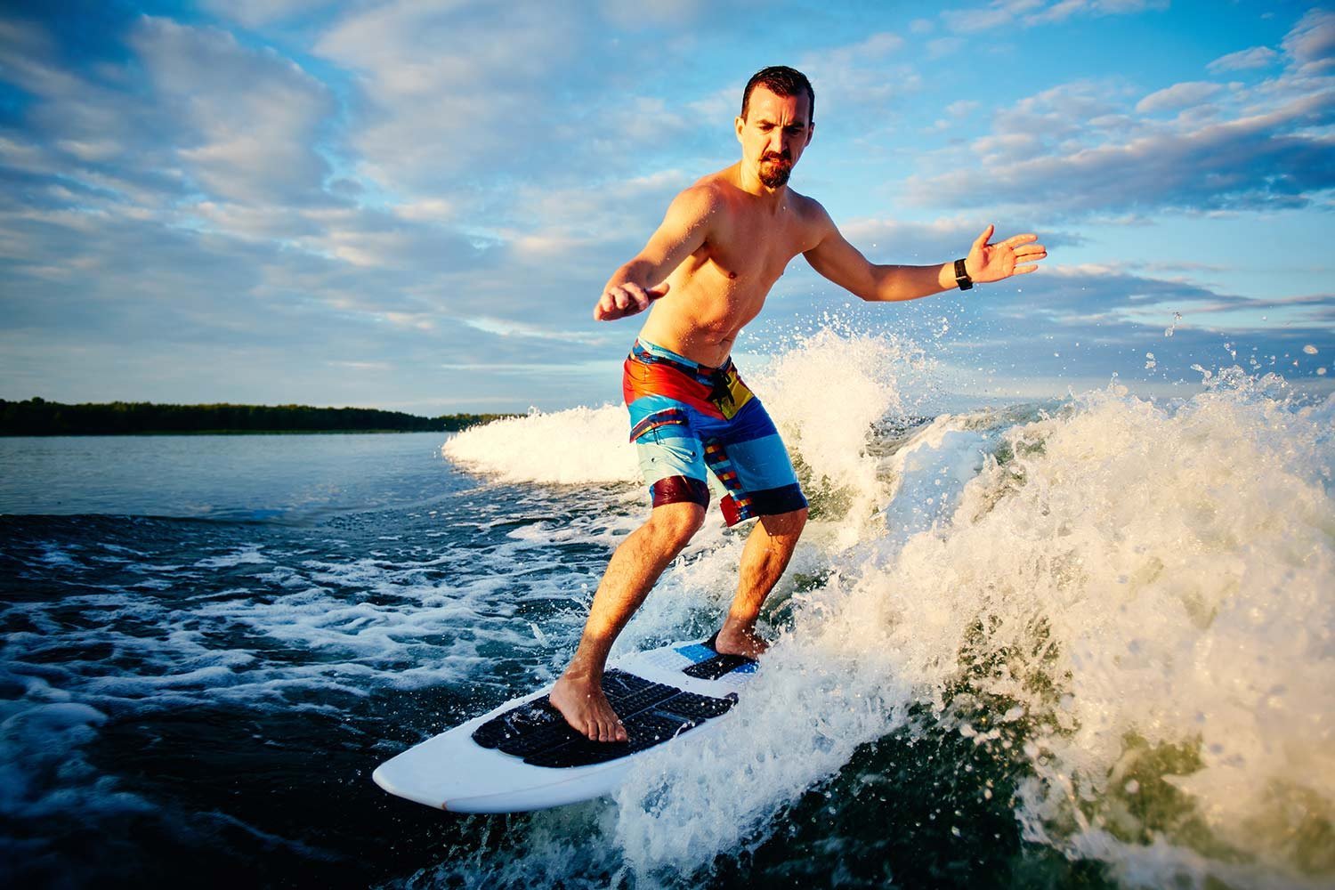 Man shirtless, wearing colorful board shorts, stands on a surfboard riding a wave with spray around him on a blue sky day