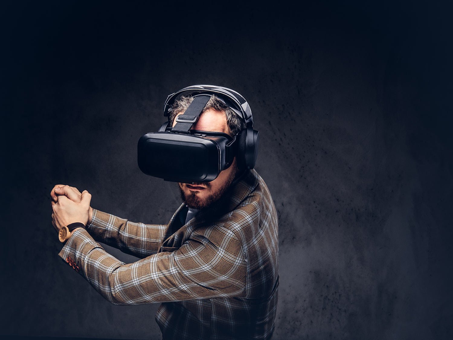 Man in a plaid blazer wearing a virtual reality headset and over-ear headphones, arms raised in a ready pose to explore VR.