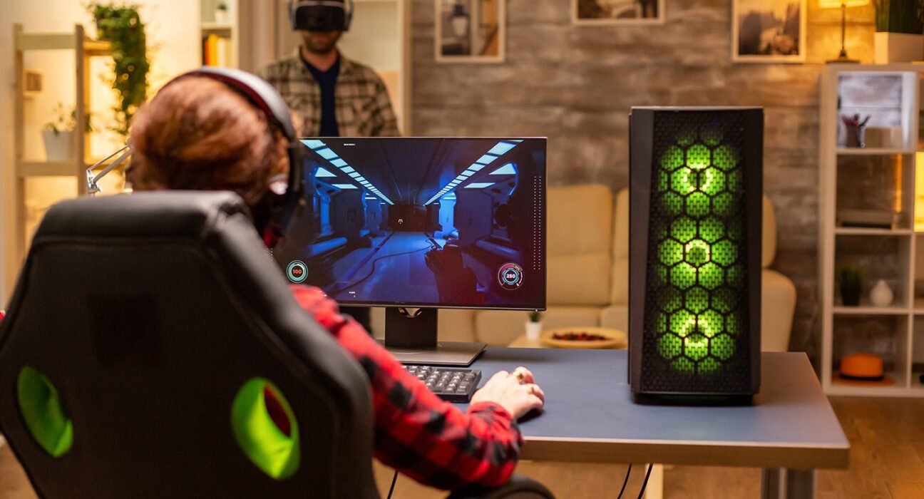 find more africa #findmoreafrica › Person in a red plaid shirt wearing a headset plays a shooter game on a desktop monitor, with a green-lit gaming PC tower on the right.
