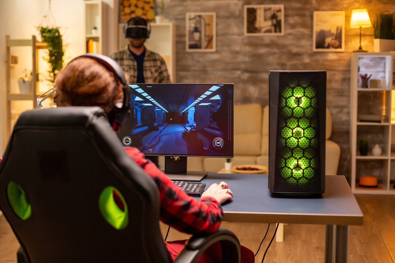 Person in a red plaid shirt wearing a headset plays a shooter game on a desktop monitor, with a green-lit gaming PC tower on the right.