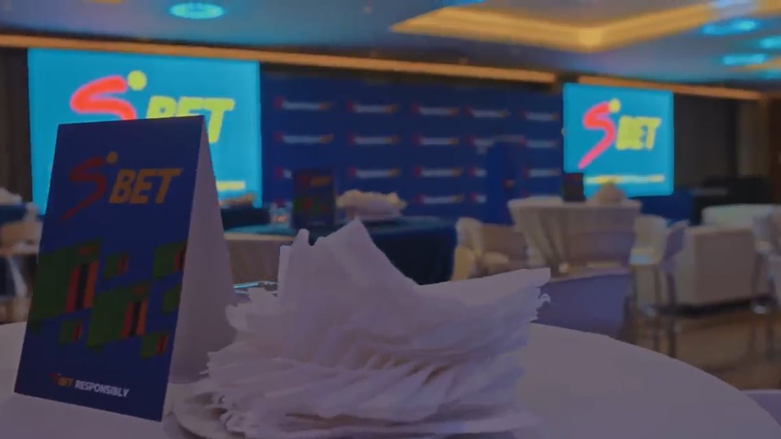 Table in a conference room with SBET branding on blue screens in the background; napkins stacked on the table in the foreground.