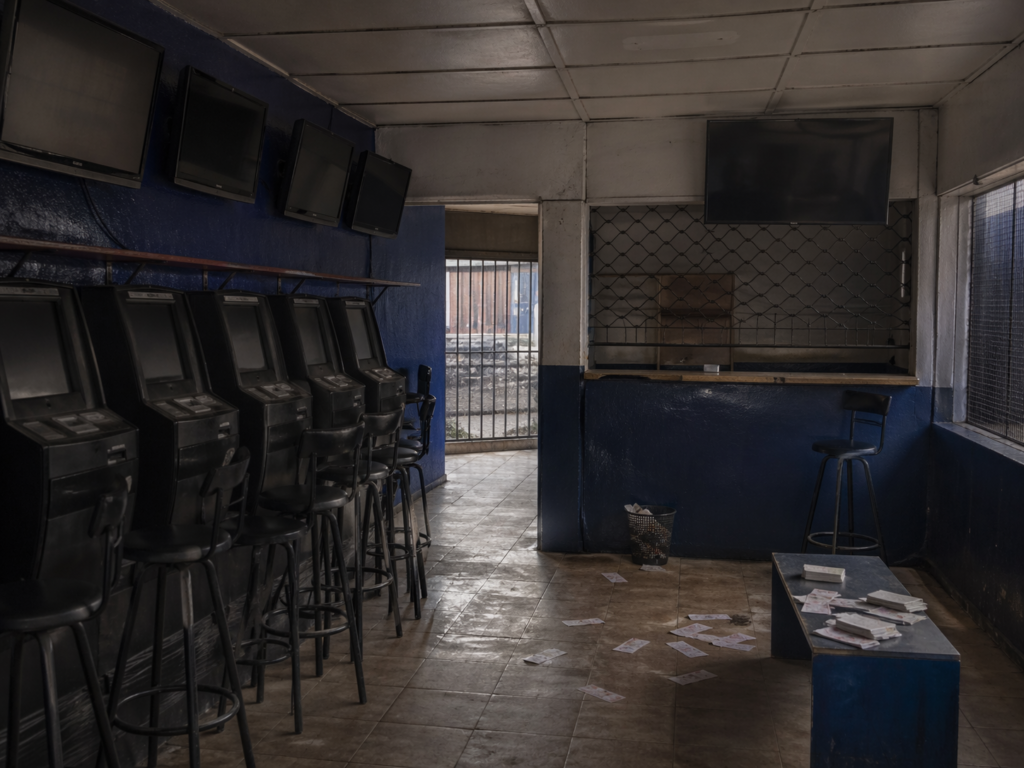find more africa #findmoreafrica › Dimly lit, empty arcade/betting shop with tall black kiosks along a blue wall and stools; papers scattered on the tiled floor.