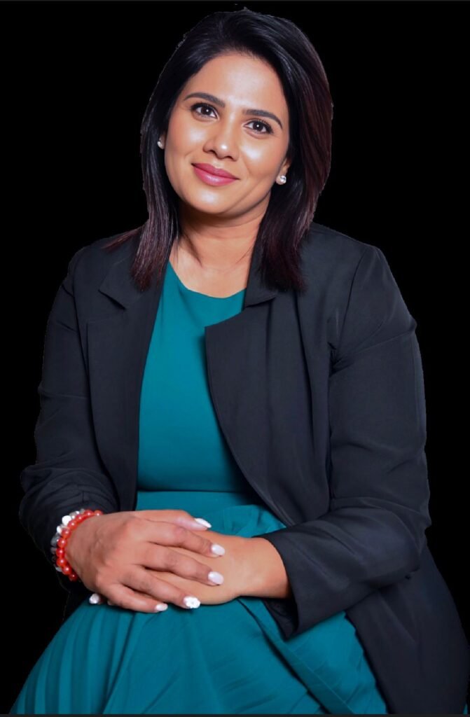 Professional woman in a teal dress and black blazer seated with hands folded, smiling at the camera against a black background.