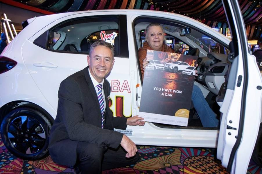 Smiling man in a suit kneels beside a white car, presenting a 'You have won a car' poster to a woman in the driver's seat at a colorful event.