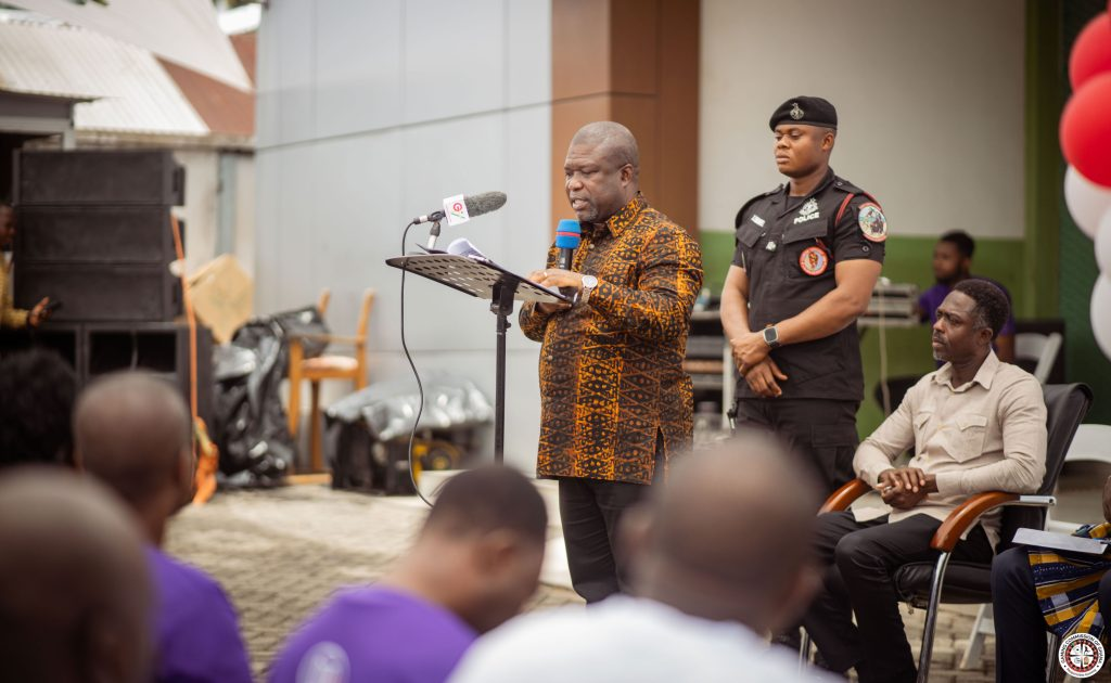 Man in a patterned shirt speaks at a podium with a microphone as a police officer stands behind and attendees listen.