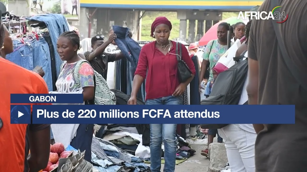 Market scene in Gabon with shoppers browsing clothes; a blue news banner reads 'Plus de 220 millions FCFA attendus' and Africa24 logo is top right.