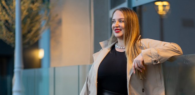 A stylish woman with long blond hair wearing a beige trench coat and black top, posing outdoors with a confident smile.