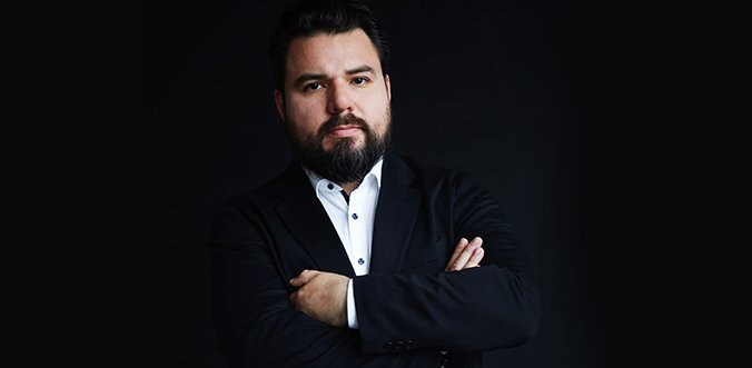 Bearded man in a dark suit with a white shirt, arms crossed in a studio portrait against a dark background.
