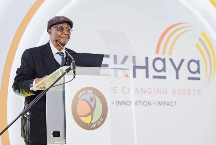 Man in a suit and flat cap delivers a speech at a podium with a microphone, in front of a backdrop showing a National Lottery logo and 'Ekhaya' branding.