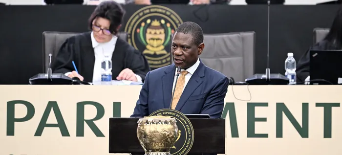 A man in a dark blue suit and gold tie speaks at a parliamentary podium; behind him are officials seated and the Parliament emblem is visible on the lectern.