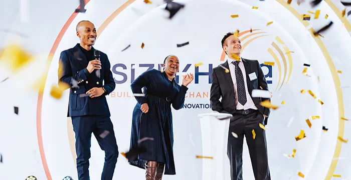 find more africa #findmoreafrica › Three adults on a stage celebrate as golden confetti falls during a conference, with a circular logo/banner in the background.