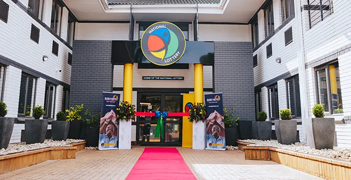 find more africa #findmoreafrica › Entrance to a National Lottery building with a red carpet, flower arrangements, and side banners reading 'National Lottery' and 'Home of the National Lottery'.