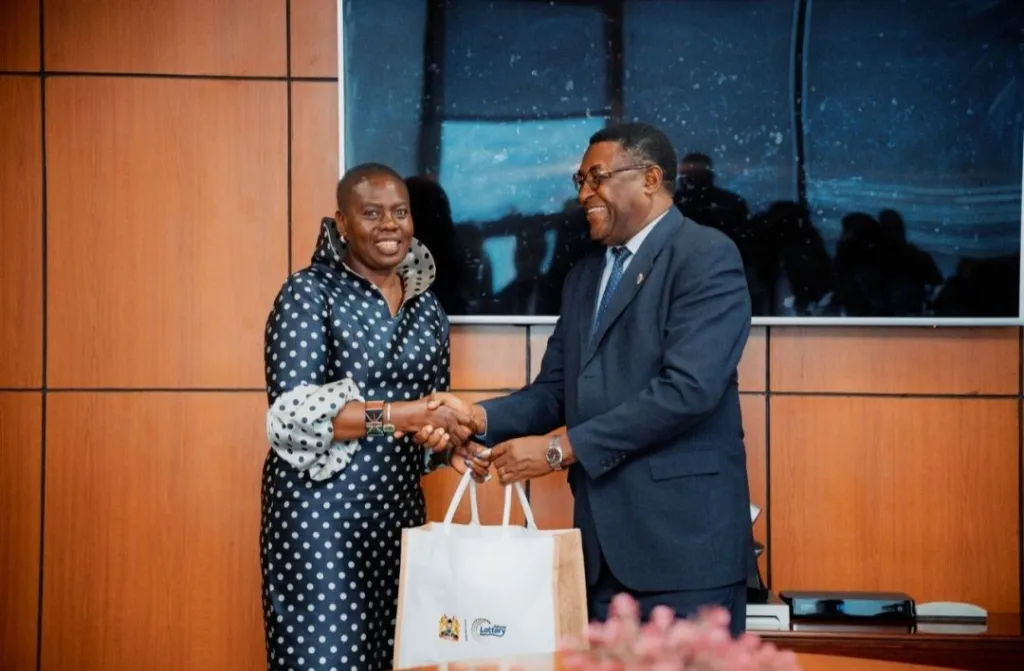 Man in a dark suit shakes hands with a woman in a polka-dot dress while presenting a beige tote bag in a wood-paneled room.