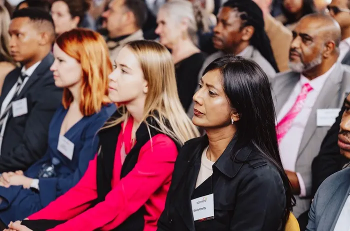 Front row of diverse conference attendees listening attentively; woman in black jacket in foreground.