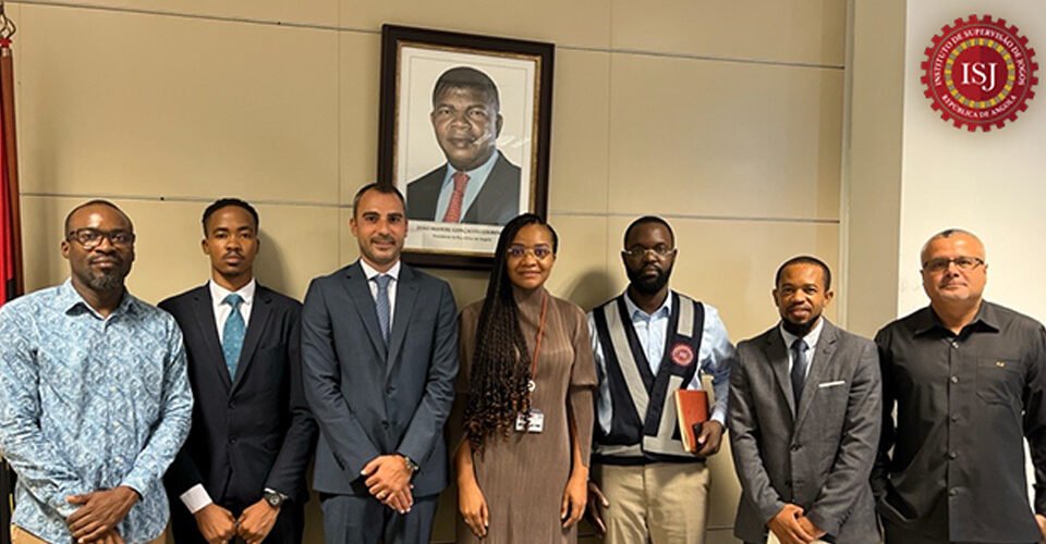 Seven professionals in business attire stand in a row in a conference room, with a framed portrait and an ISJ emblem in the background.