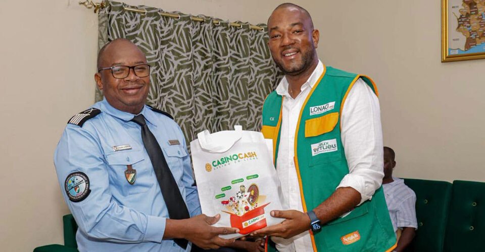 Two men exchange a white bag labeled 'CASINO CASH' while smiling; one wears a light blue security/uniform shirt, the other a green-yellow vest in a room with a patterned curtain.